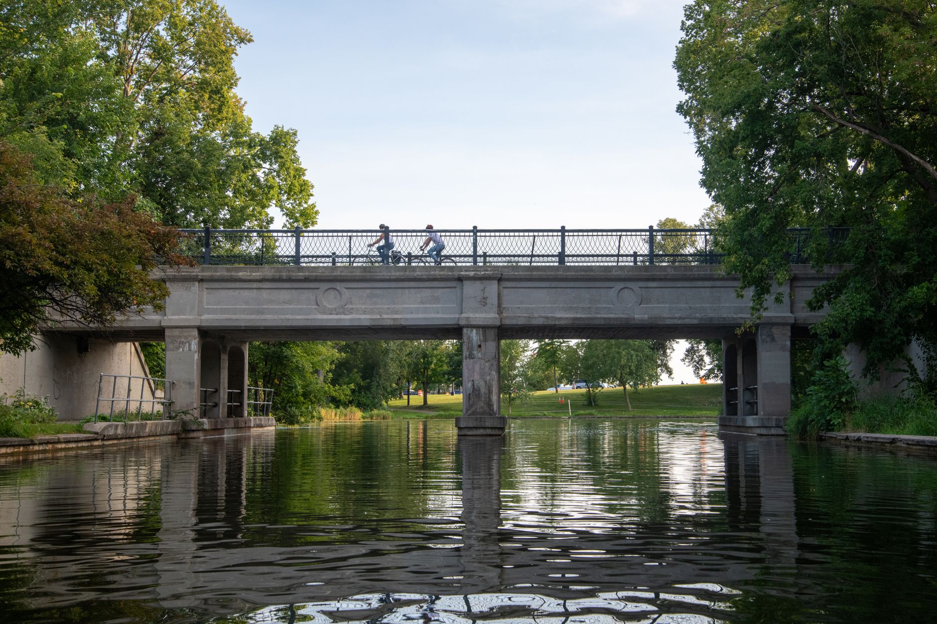 Cyclists cross a historic stone bridge over calm green parkland water